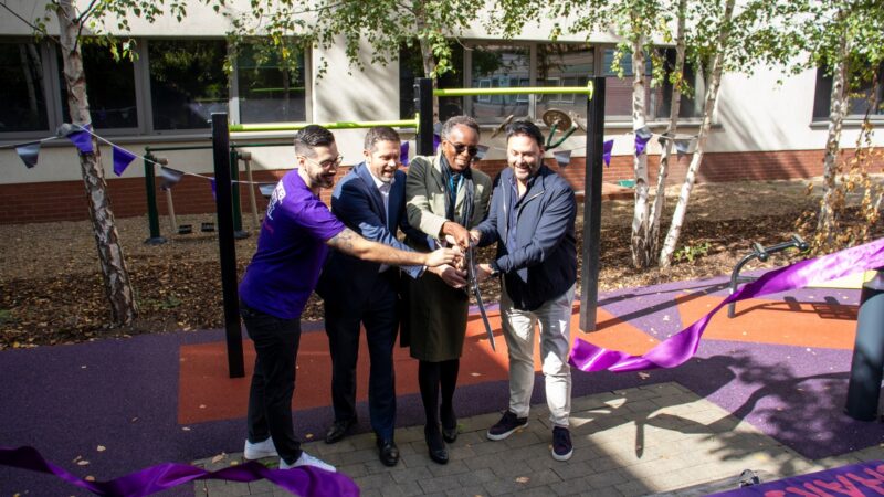Four people standing in a row holding large scissors and cutting a ribbon in front of some outdoor gym equipment.