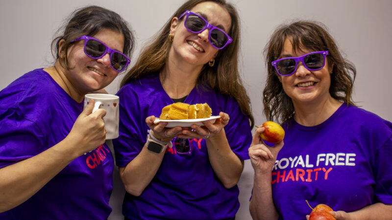 Three Royal Free Charity supporters wearing purple T-shirts and glasses pose with cake and tea at a tea party fundraiser.