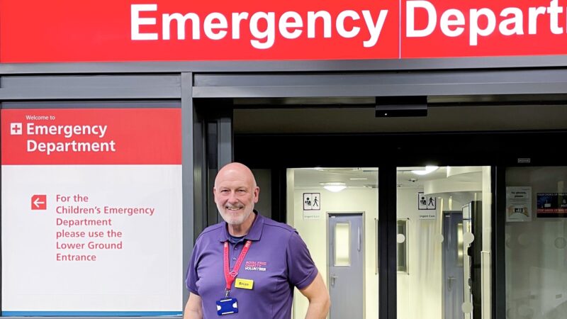 Brian wears a purple volunteer polo shirt. He is standing in front of the entrance to an emergency department.