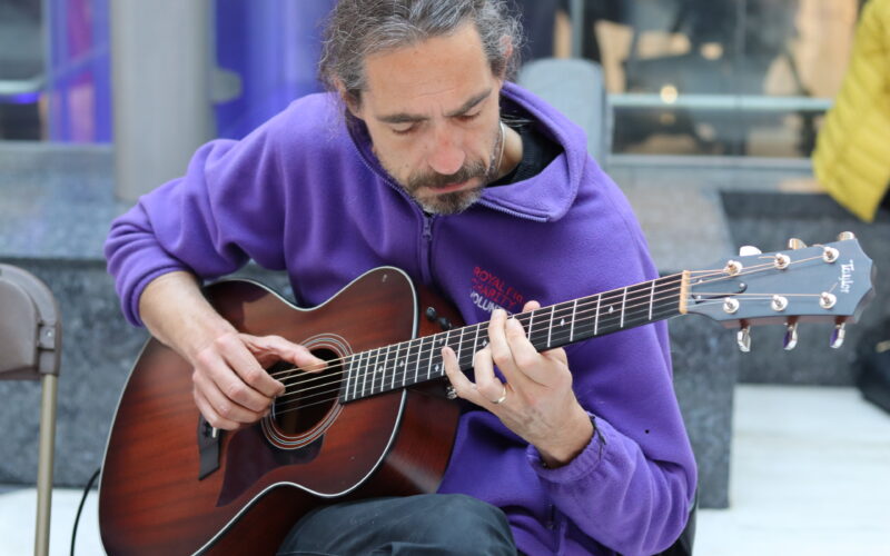 Man playing acoustic guitar, sat on a chair.