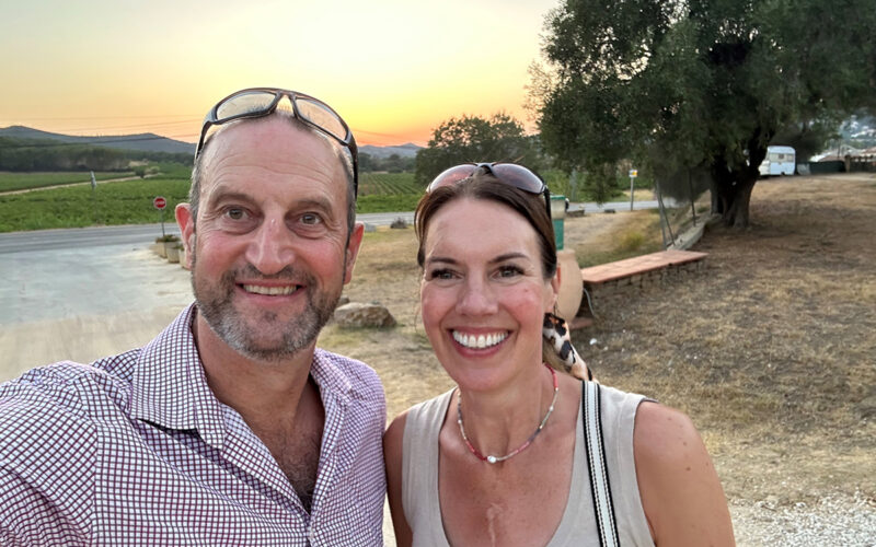 A man and woman with their arms around each other standing in front of a tree by a river under the setting sun.