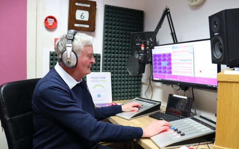 Man sat at a radio desk wearing headphones and speaking into a mic.
