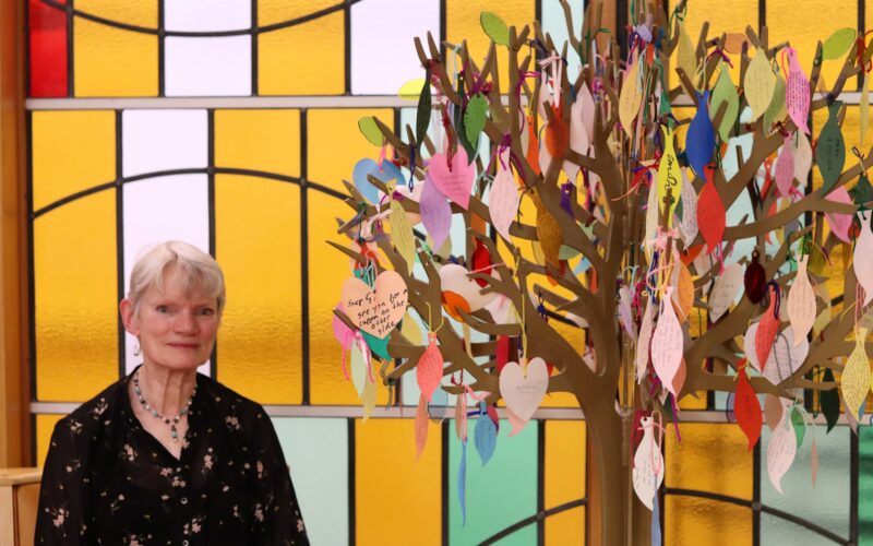 A woman sat next to a craft tree covered in small pieces of colourful paper to resemble leaves on which prayers are written.
