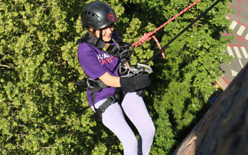 A woman wearing a Royal Free Charity t-shirt, abseiling down the Pears Building. The background is green leaves on a tree.