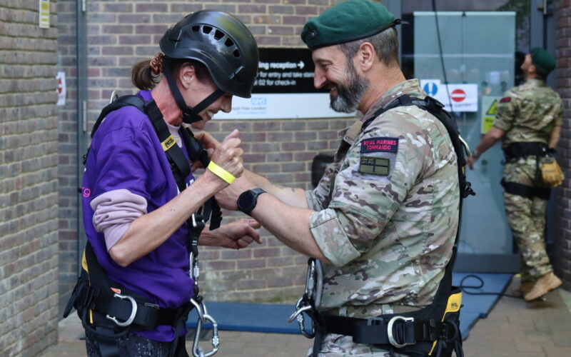 A woman wearing a harness and helmet stood next to a male Royal Marine who is helping her take off her helmet.