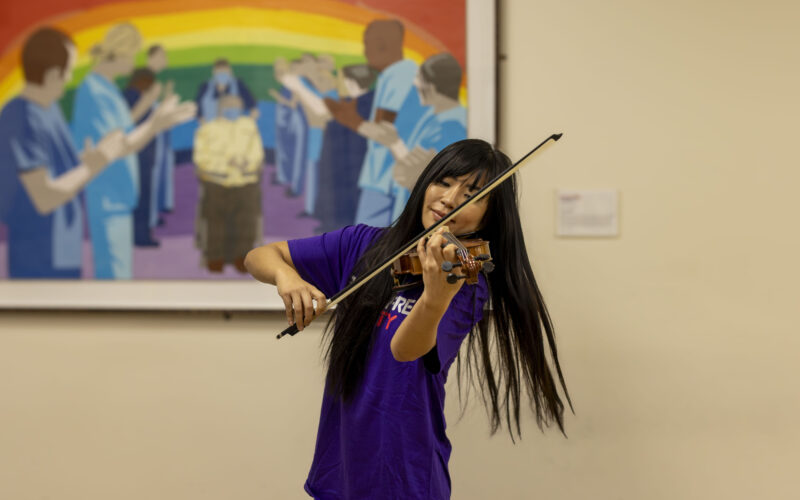 A woman playing violin with her eyes closed. She is wearing a Royal Free Charity t-shirt and standing in a hospital corridor with a piece of art hanging on the wall behind her.