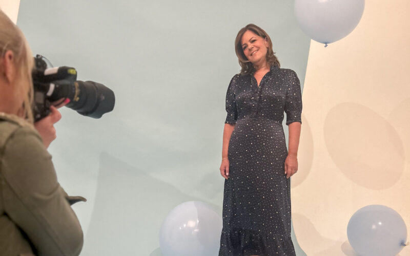 A woman is posing for a photo shoot. The photographer can be seen to the side of the shot.