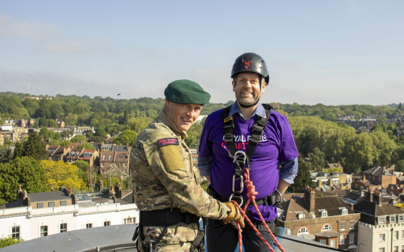 A man wearing a Royal Free Charity tshirt preparing to abseil. He is wearing a harness and helmet and he is stood by a male Royal Marine.