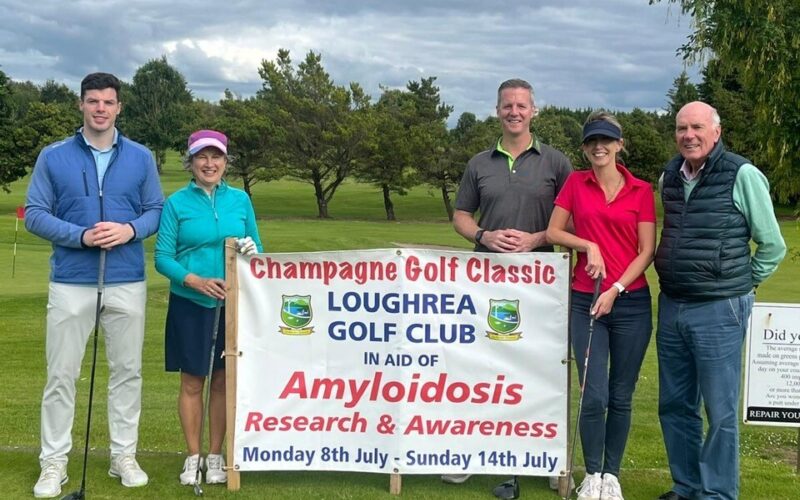 A group of golfers standing each side of a sign that reads "Champagne Golf Classic, Loughrea Golf Club in aid of Amyloidosis Research and Awareness - Monday 8th July  - Sunday 14th July."