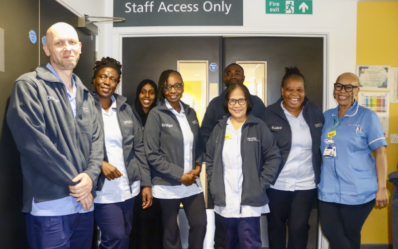 A group of hospital staff members, dressed in light blue uniforms and grey fleece jackets, stand together in front of a door labeled "Staff Access Only." Some staff have name tags or embroidered names on their jackets, and the setting is a hospital corridor with signage and informational posters visible.