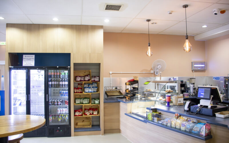 A hospital coffee shop area with a table, till and fridge.