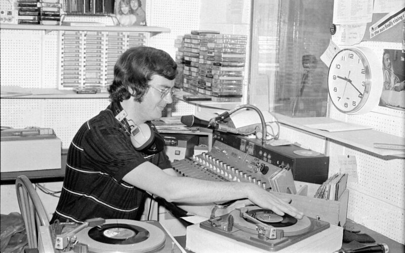 A black and white photo of a man sat at a radio desk surrounded by tapes. The clock on the wall reads 9:20am.