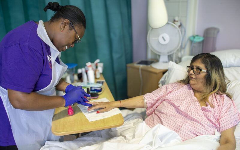 A female beautician wearing a Royal Free Charity t-shirt painting the nails of a female patient who is lying in a hospital bed - both are smiling.