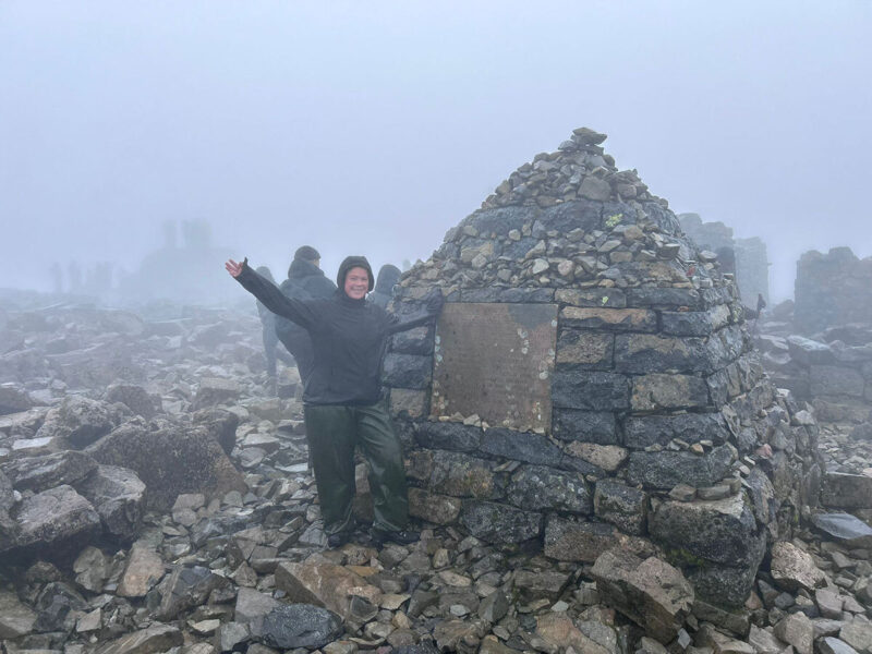 A woman in outdoor trekking grear standing beside a small hut made of bricks and rocks on the top of a misty mountain.