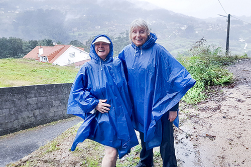 Two women stood outside together in front of a country lane, wearing waterproofs.