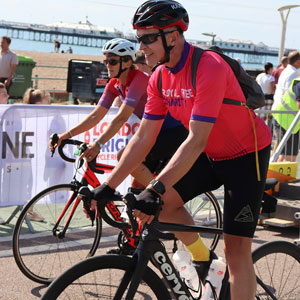 Two people in Royal Free Charity cycling shirts riding along Brighton Pier in the day time with others. 