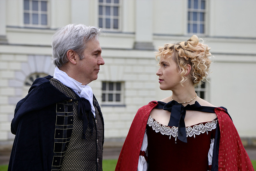 A man and women in period drama costume, outside in front of a stately manor.