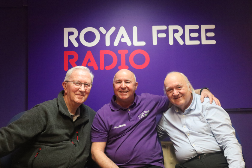 Three men sat with their arms around each other in front of a sign that says Royal Free Radio.