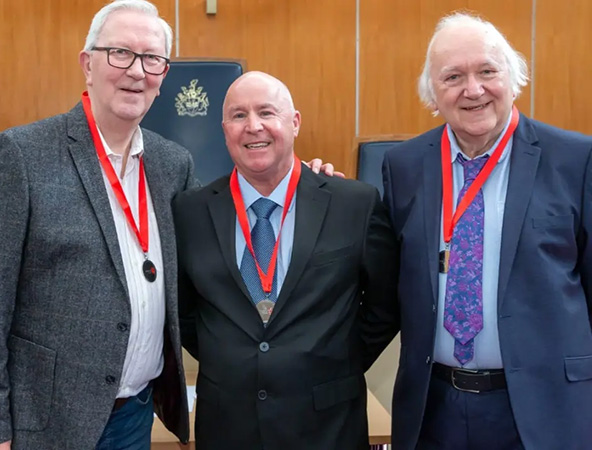 Three men in smart attire, standing in a row, each of them wearing a medal. 